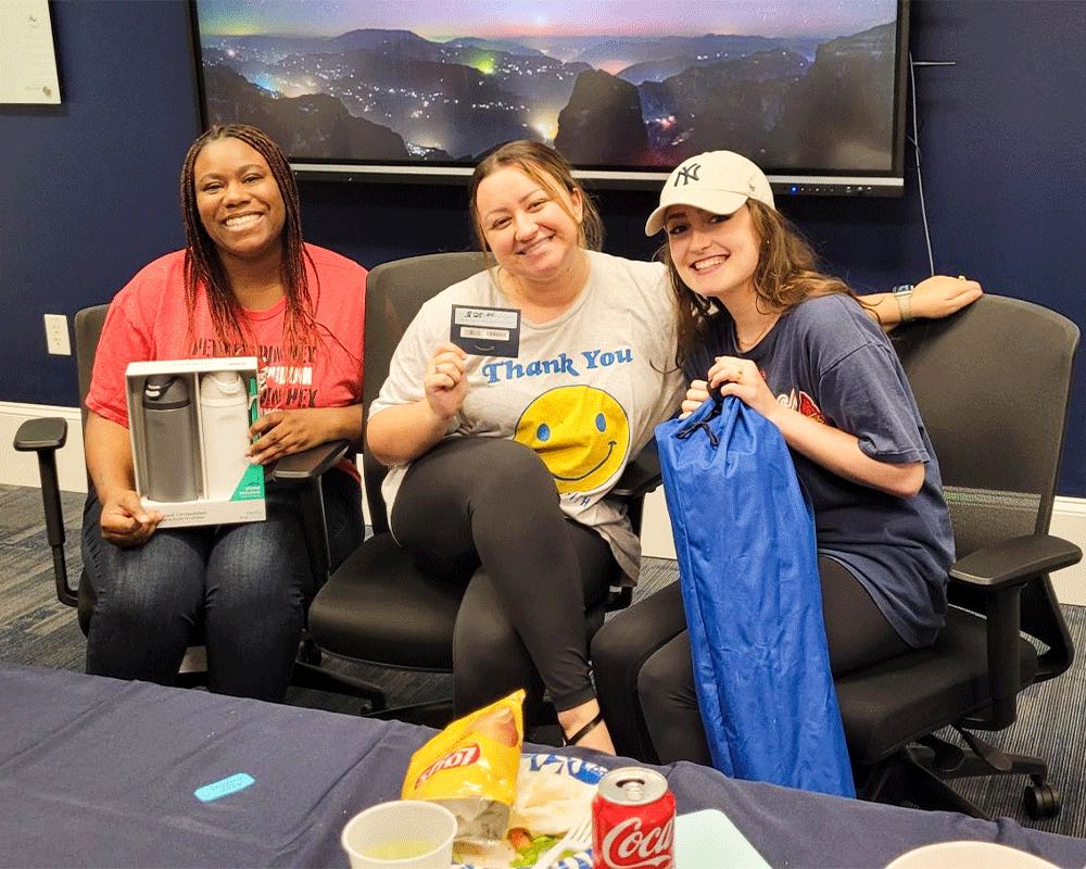 Three coworkers smile in office chairs holding prizes: a water bottle set, a gift card and a blue camp chair bag.