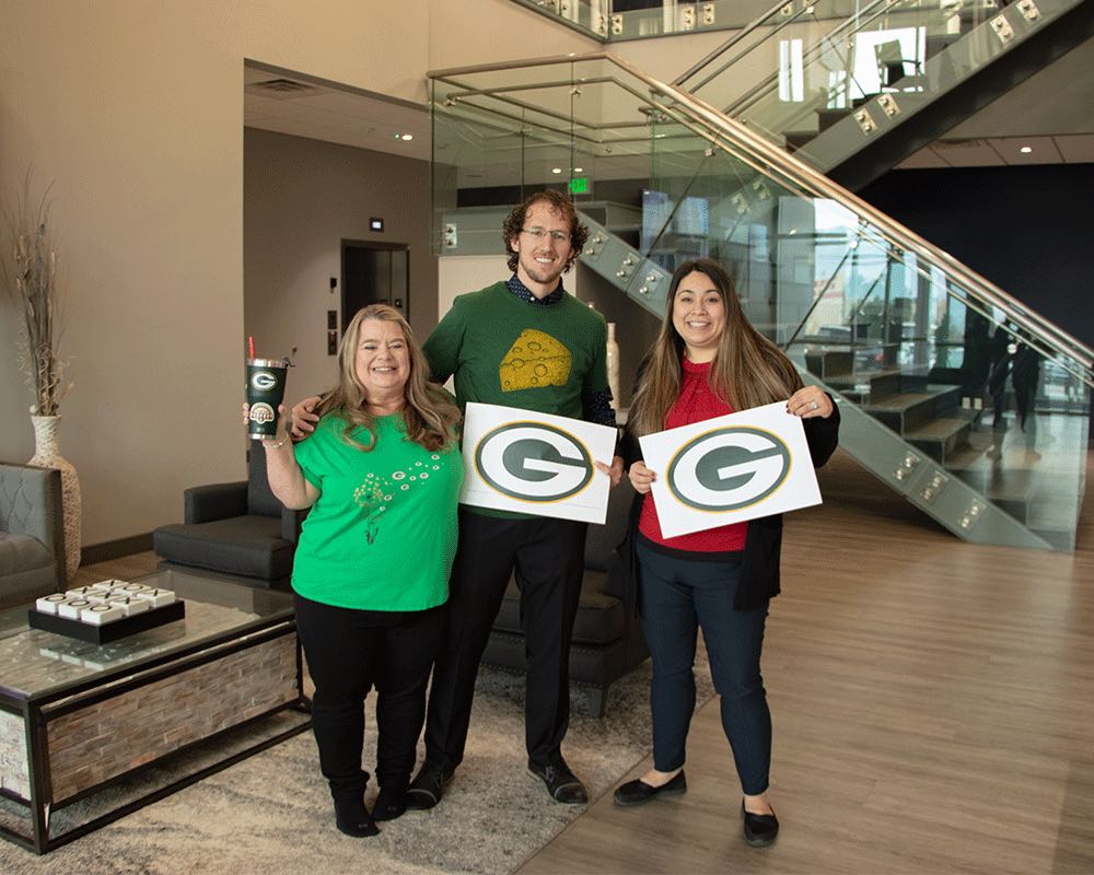 Zack, Sam and JoBeth in the office lobby in green, holding Green Bay Packers signs.