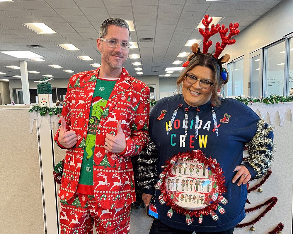 Two coworkers in holiday outfits, one in a red reindeer suit and one in a Holiday Crew sweater and antlers.