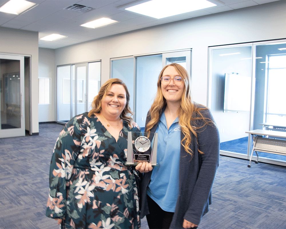 Two coworkers in an office hallway, one holding a work anniversary award.