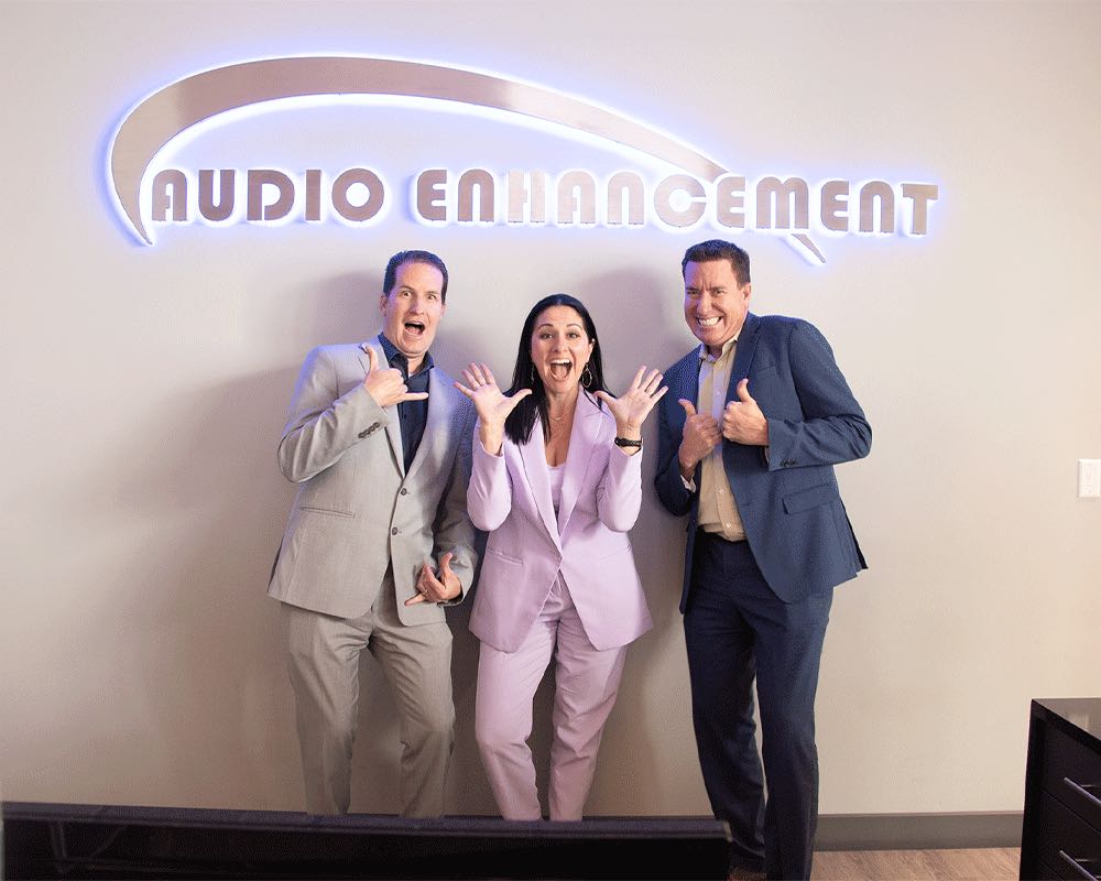 Three coworkers make playful poses under the lit Audio Enhancement sign.