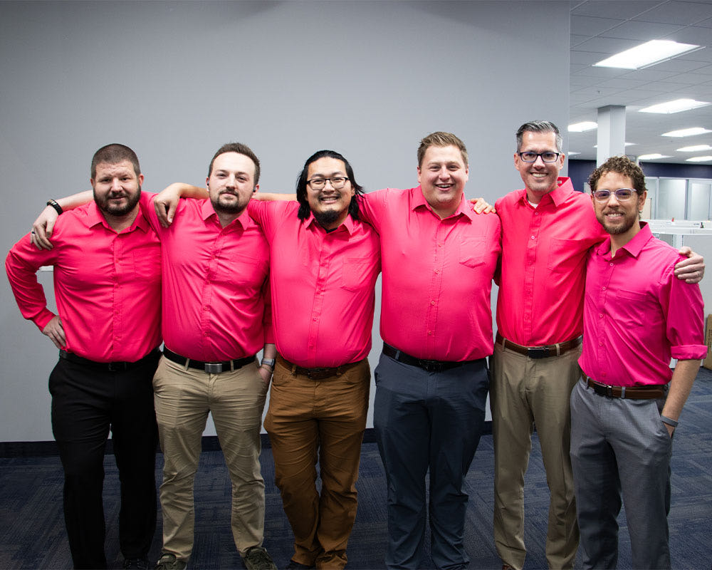 Six coworkers in matching pink shirts stand arm in arm.